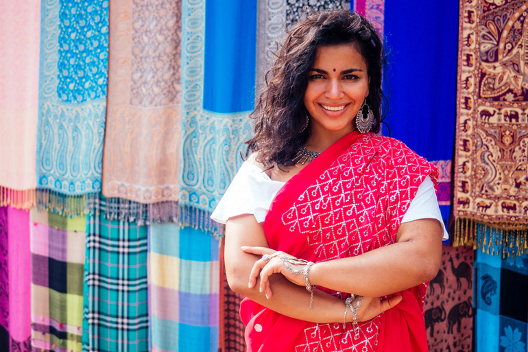 Une femme souriante en sari pose devant des tissus colorés suspendus.