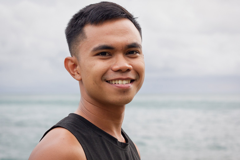 Un jeune homme au sourire serein pose de profil en extérieur devant une étendue d’eau calme.