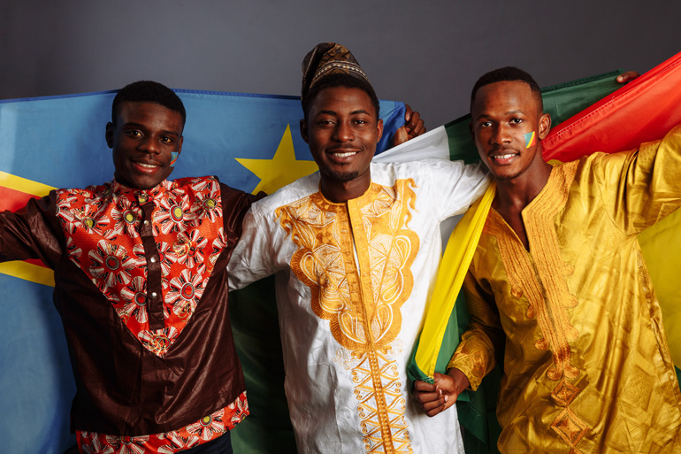 Trois jeunes hommes souriants en tuniques traditionnelles colorées posent avec des drapeaux africains.