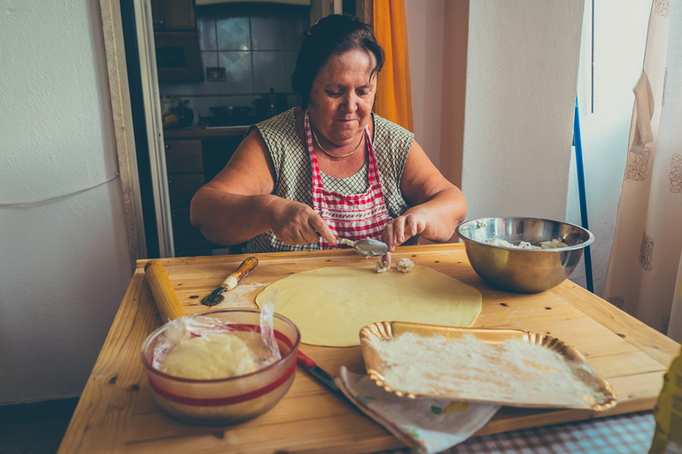 Une femme âgée portant un tablier prépare avec attention des pâtes sur une table en bois.
