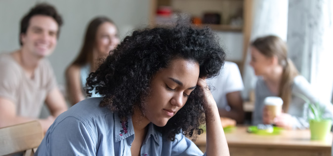 Une jeune fille triste a la tête baissée sur une table pendant que d’autres ados sourient derrière elle.